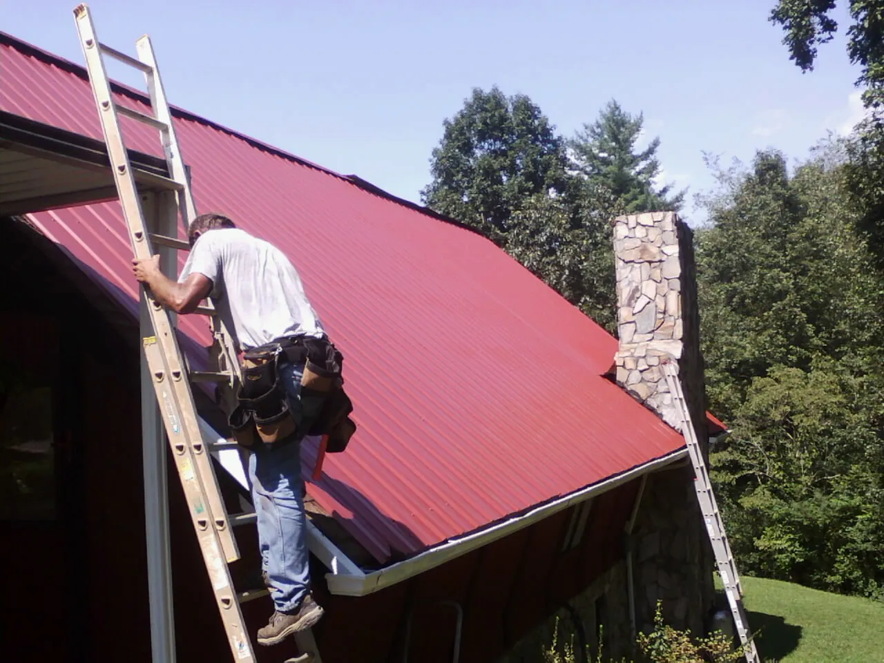 Roofer on a ladder beside a red metal roof and stone chimney — Soffit & Fascia Repair by Roofing Brothers in Scotts Hill